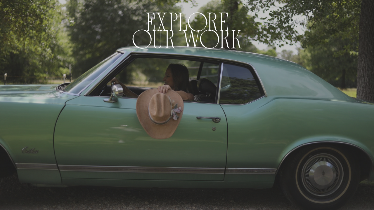 Vintage green car with a person inside and a hat on the dashboard, surrounded by trees.